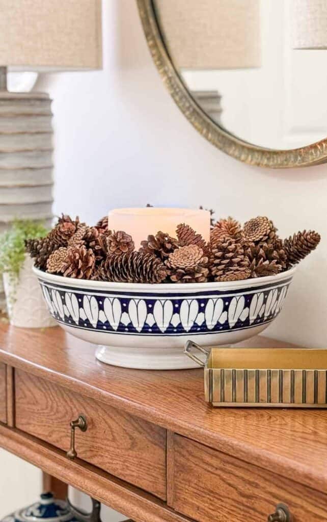 Wood entryway console styled with a blue and white patterned bowl filled with pinecones and a glowing candle, a brass matchbox, and a ribbed white lamp beneath a round gold mirror.