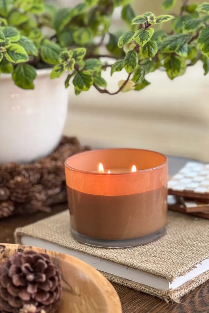 A lit fall candle in an amber glass jar sitting on a burlap covered book beside a pinecone.