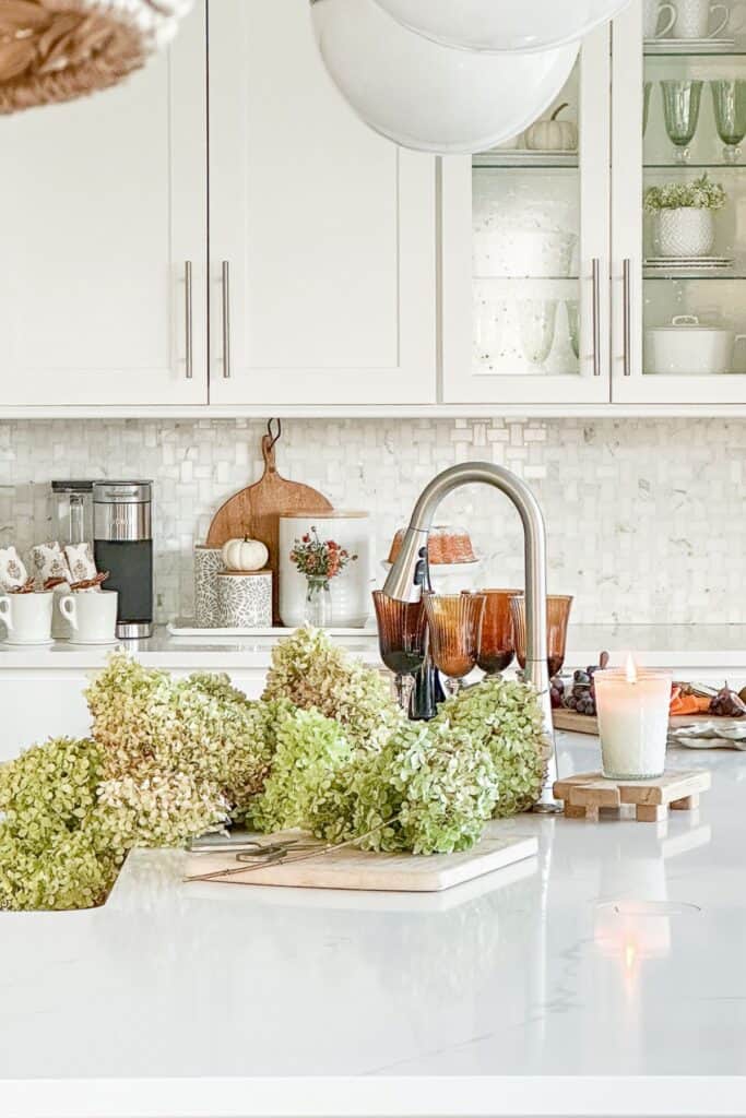 White kitchen with a fall coffee station, amber glassware, a lit candle, and fresh cut hydrangeas on the island