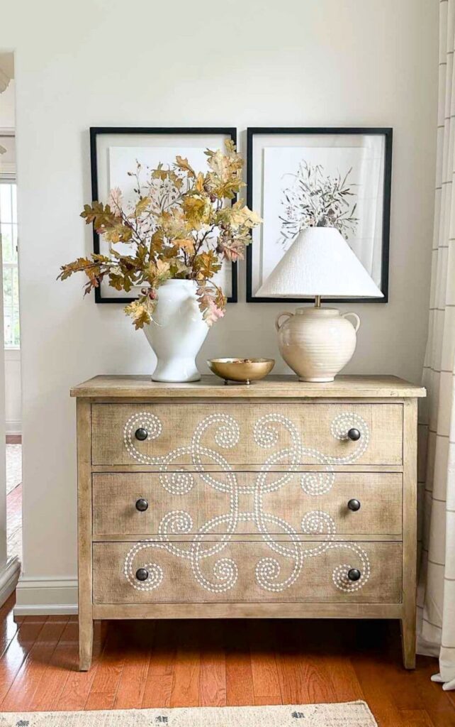 Fall leaf branches in a white vase on a wood dresser with framed art and a table lamp