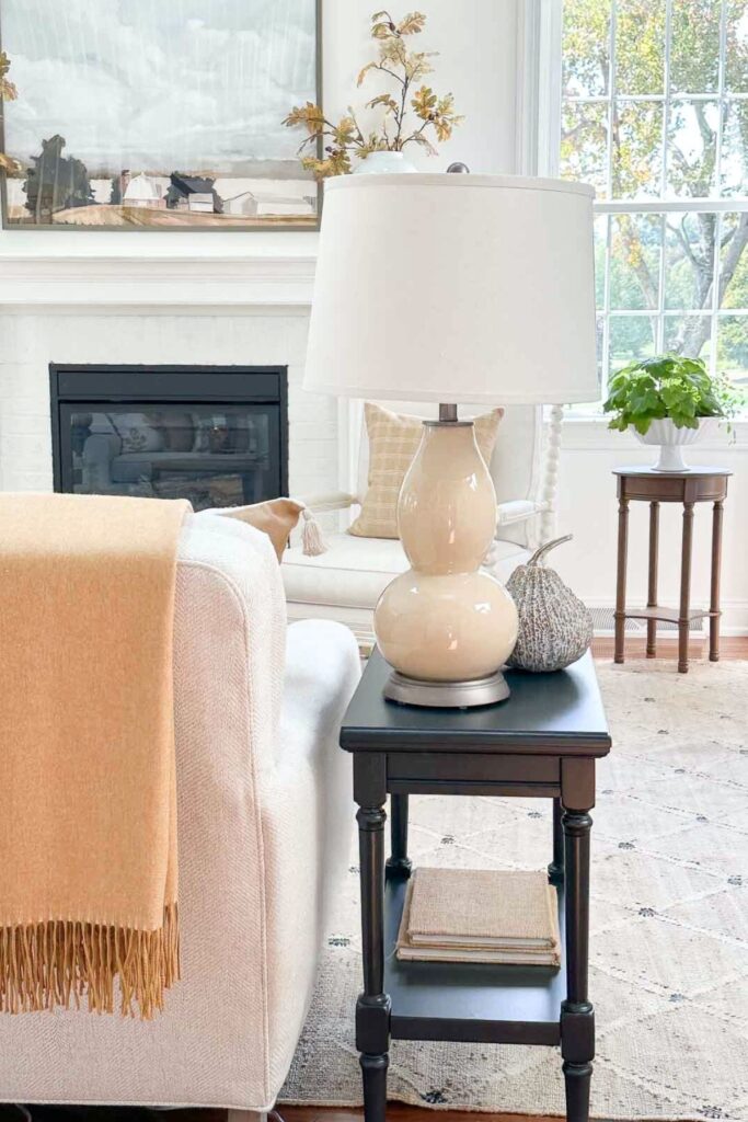 A mustard fall throw draped over a sofa arm beside a black side table styled with a lamp and mini pumpkin, in a cozy fall living room.