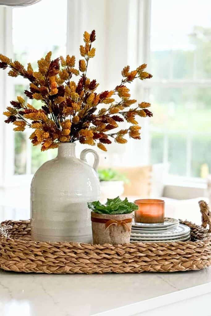 Fall vignette on a kitchen island with dried autumn stems in a white jug, small plant, candle, and plates on a woven tray