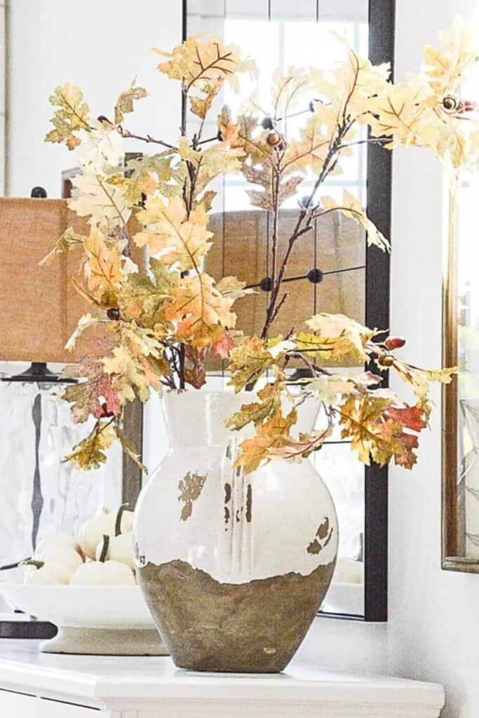 Tall faux fall leaf branches in a white and stone vase on a mantel with white pumpkins nearby