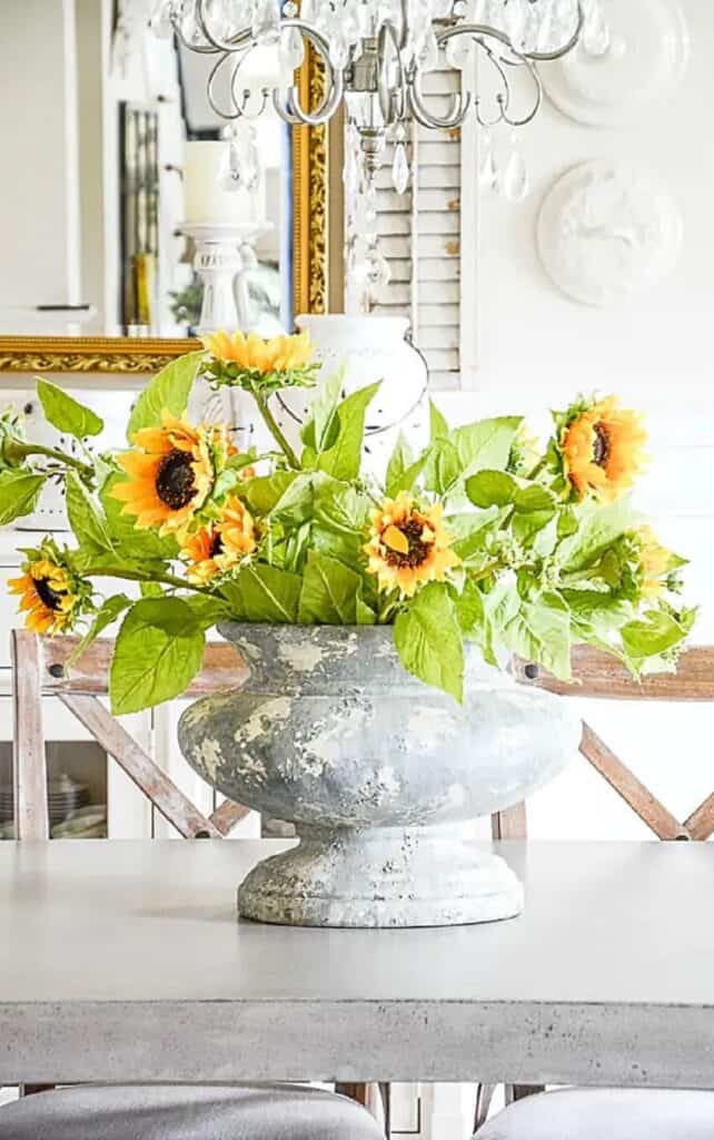 Faux sunflowers in a weathered stone urn on a dining room table beneath a crystal chandelier