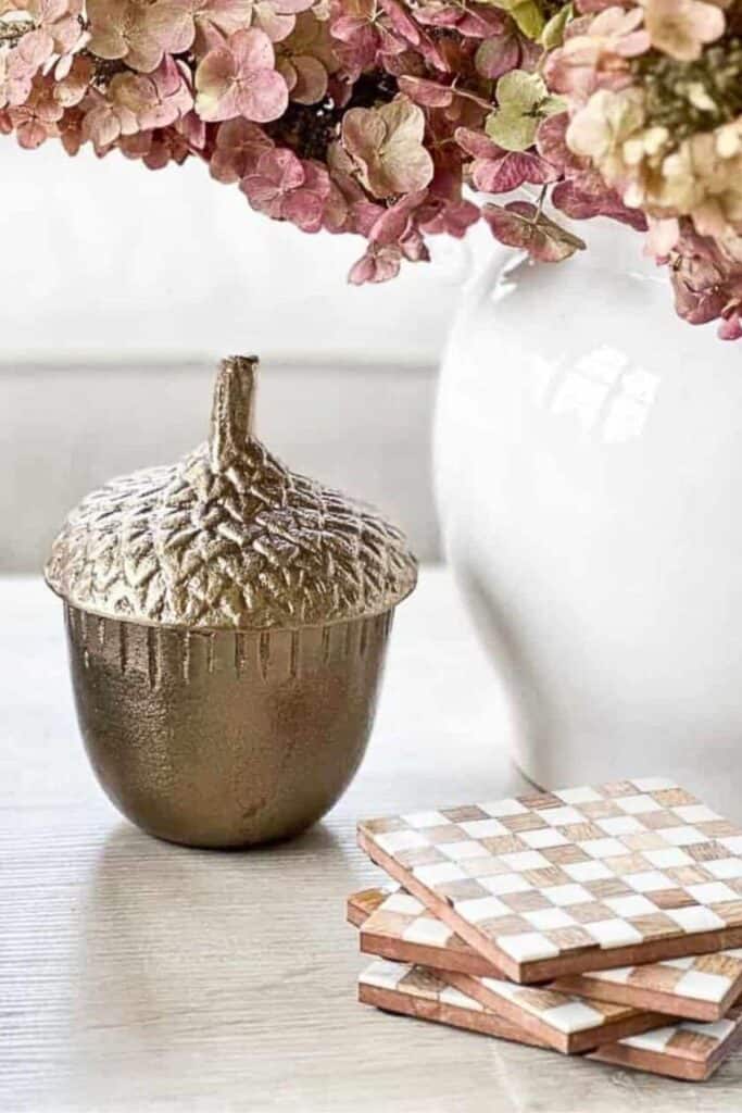 Gold acorn box with dried pink hydrangeas and checkered coasters on a table, a subtle early fall decorating touch