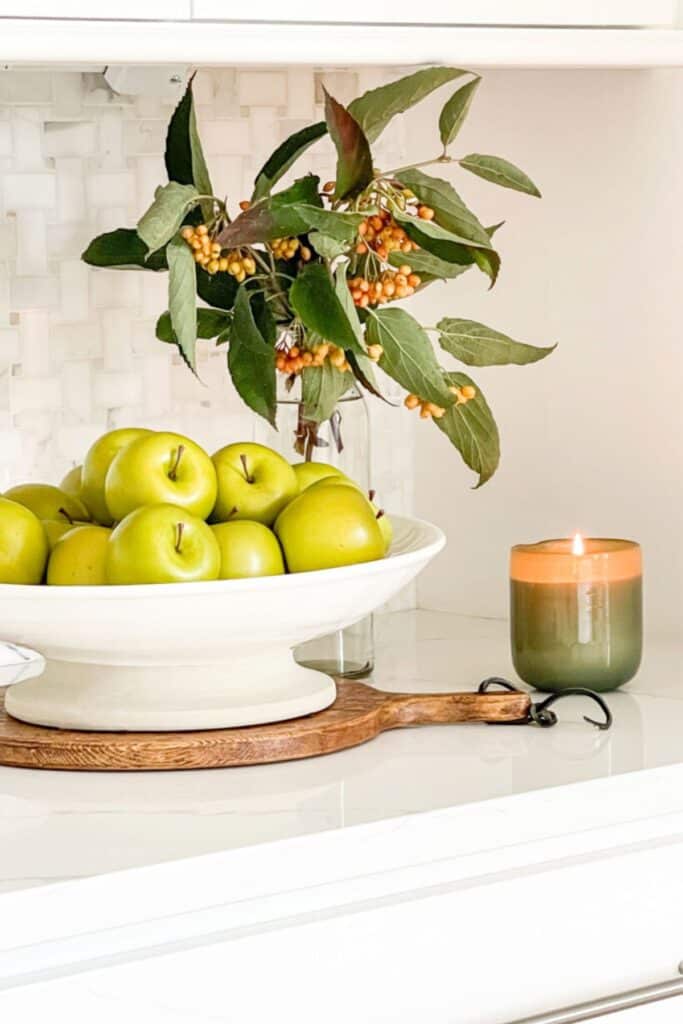 White pedestal bowl of green apples on a wood board with a lit candle and foraged berry branch on a kitchen counter