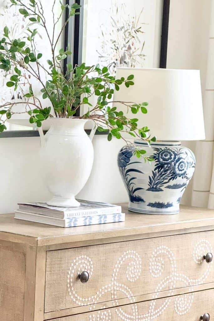 Wooden dresser with stenciled swirl design styled with a blue and white floral lamp, a white pitcher of greenery, a small glass cloche, and a stack of books beneath framed botanical art.