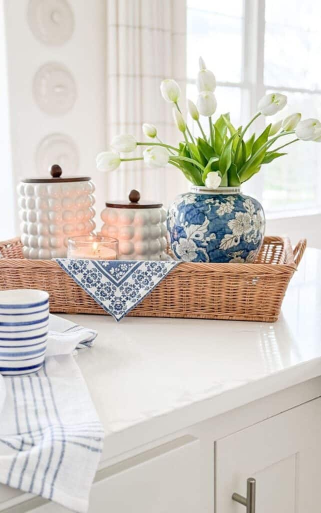 Kitchen island styled with a woven tray holding white hobnail canisters, a lit candle, a blue floral linen napkin, and a blue and white vase filled with white tulips.