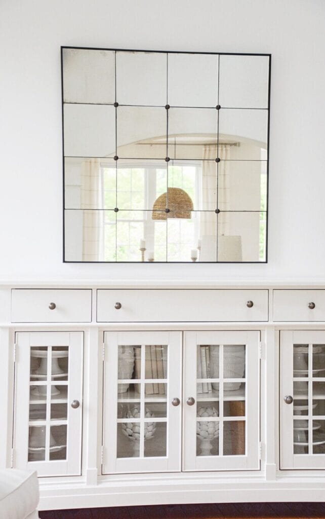 Large paned mirror with black grid frame hung above a white built in cabinet with glass front doors, styled with cake stands, vases, and stacked books.