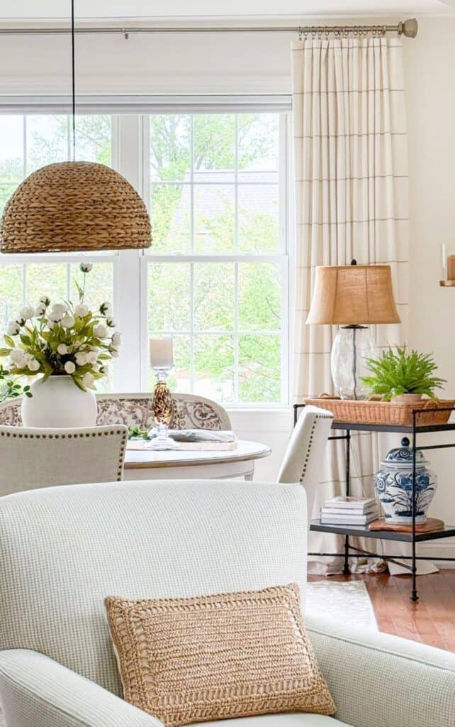 Open living and dining area with a woven pendant light, round dining table styled with white florals, and a black iron side table layered with a lamp, ginger jar, books, and a woven tray.