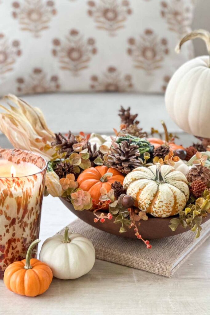 A wooden bowl filled with white and orange pumpkins, pinecones, and dried florals beside a glowing candle, styled for budget friendly fall decorating.