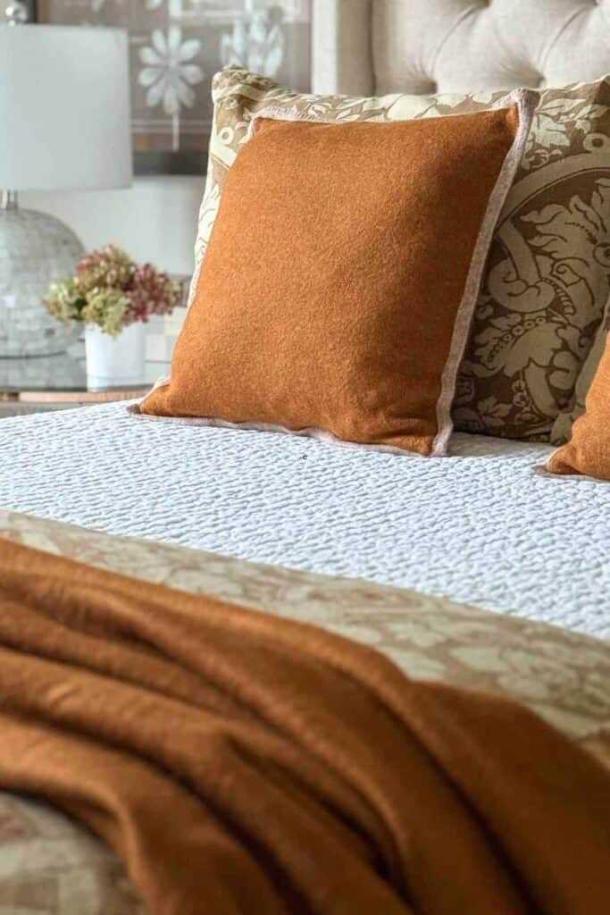 Rust colored pillow and throw layered on a textured white quilt with a patterned floral pillow and dried hydrangeas nearby