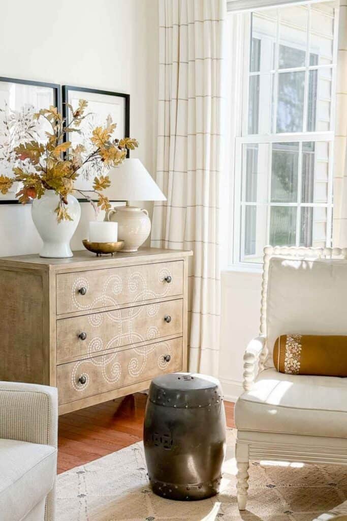 Scrolled wood chest styled with a white vase of fall leaves, a candle, and a lamp beside a cream armchair with a mustard pillow