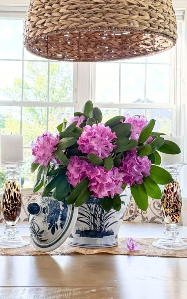 Dining table centerpiece with pink rhododendron blooms in a blue and white ginger jar, flanked by tortoiseshell glass candlesticks beneath a woven pendant light.