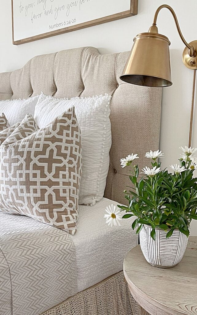 Tufted upholstered headboard layered with white quilted pillows and a taupe geometric pillow, beside a nightstand with a brass swing arm sconce and a striped planter of white daisies.