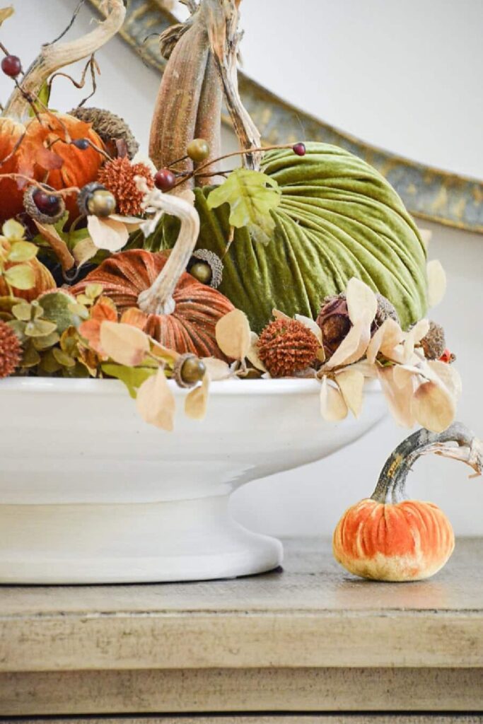 Velvet pumpkins, acorns, and dried leaves arranged in a white pedestal bowl for a budget friendly fall centerpiece.