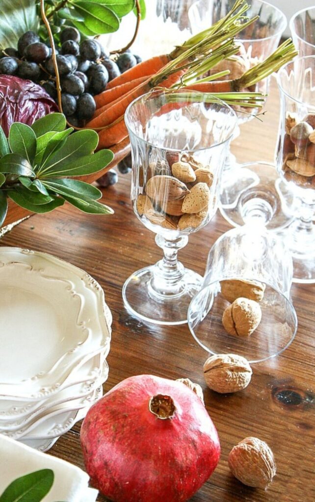 Walnuts and almonds in glass goblets with a pomegranate, carrots, and grapes on a wood table
