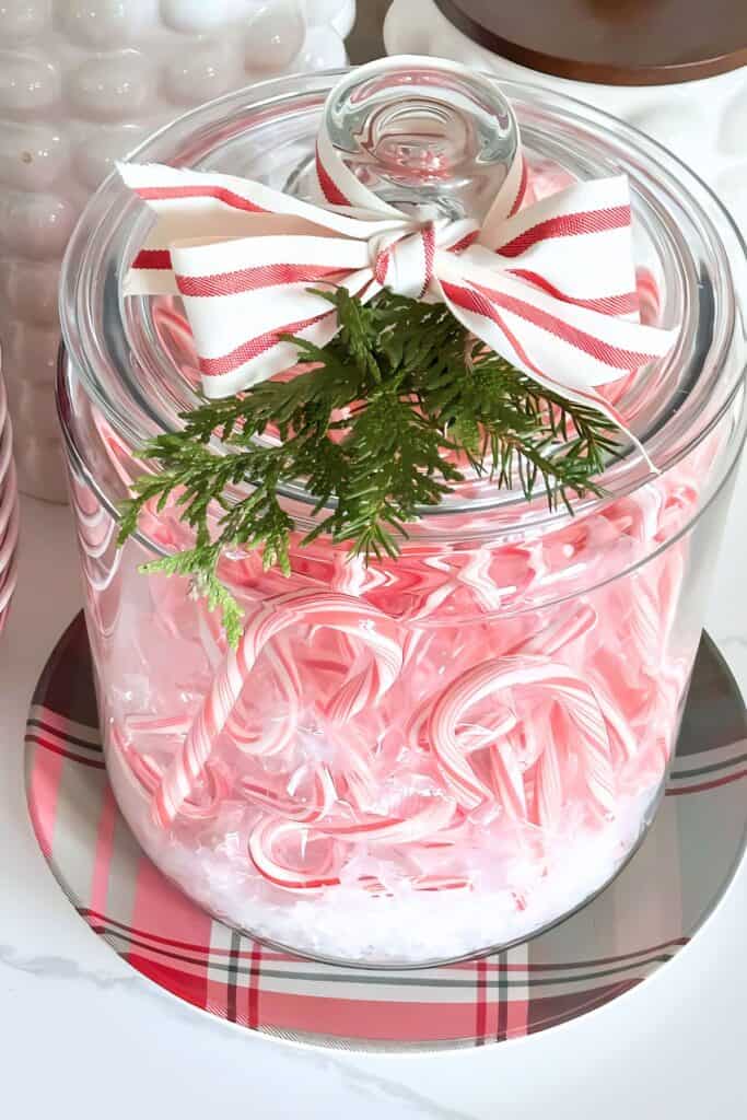 Glass jar filled with candy canes tied with a white and red striped ribbon bow and greenery — a festive Christmas 2025 decorating idea featuring bows.