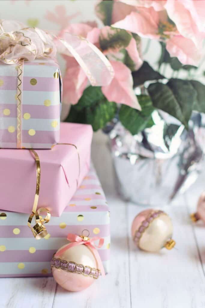 Stacked pink and white Christmas presents with gold ribbon beside pink ornaments and blush poinsettias, showing the blush pink Christmas decorating trend for 2025.