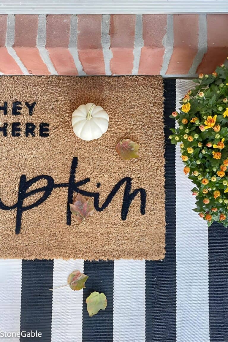 White pumpkin on a fall doormat with mums and autumn leaves by a front door — simple and welcoming pumpkin decorating idea for fall.