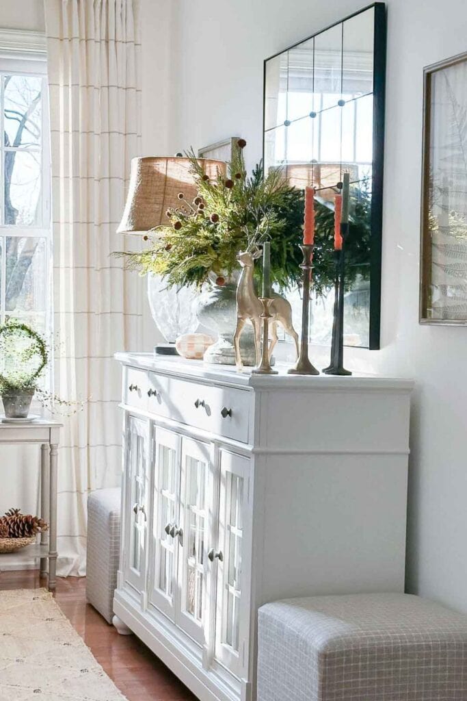 A white sideboard decorated for Christmas with natural greenery, gold deer figurines, and amber candles, showing the earthy green holiday color trend for 2025.