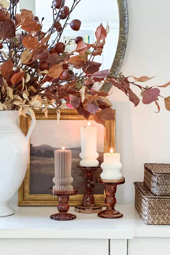 Fall decorating without orange featuring brown branches in a white pitcher, ribbed candles, and woven baskets on a white buffet.