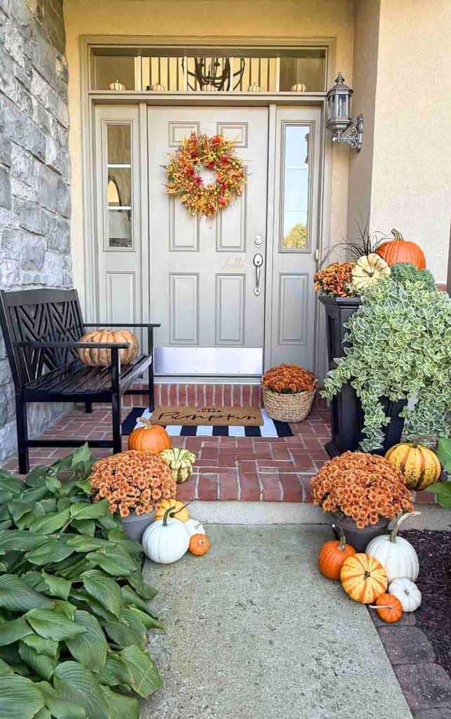 Fall front porch with orange, white, and striped pumpkins, orange mums, a planter with kale and trailing greenery, a pumpkin on a black bench, and mini pumpkins lining the door transom.