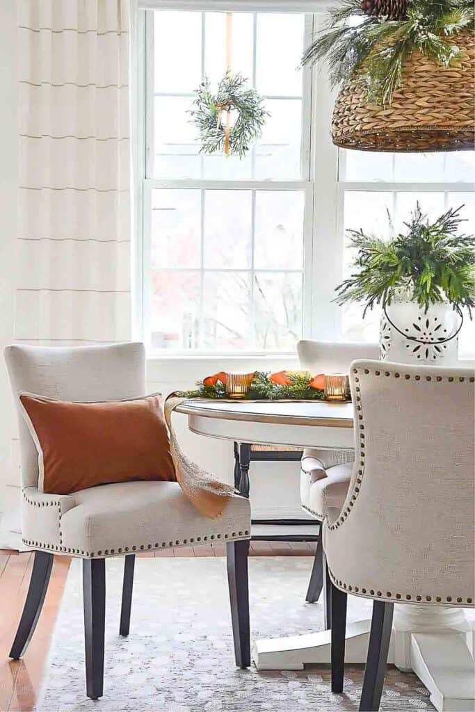 Neutral Christmas breakfast room with greenery centerpiece, caramel pillow, and woven pendant featuring pinecones and evergreens.