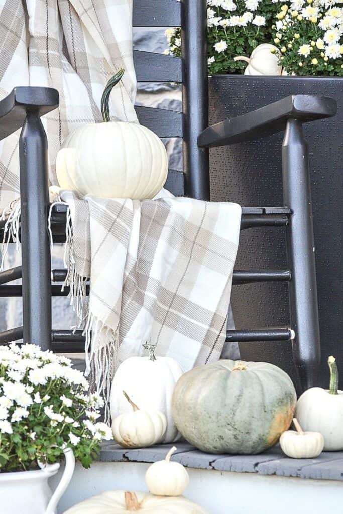 White and green pumpkins styled on a fall porch with black rocking chairs, white mums, and a neutral plaid throw blanket.