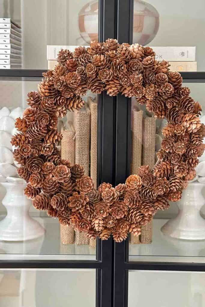 A natural pinecone wreath with warm brown tones, hanging on glass cabinet doors, representing the 2025 earthy neutral Christmas decorating trend.