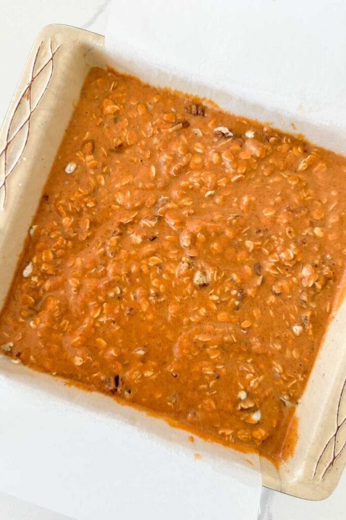 Pumpkin baked oatmeal batter with oats and pecans spread in a parchment-lined square baking dish before baking