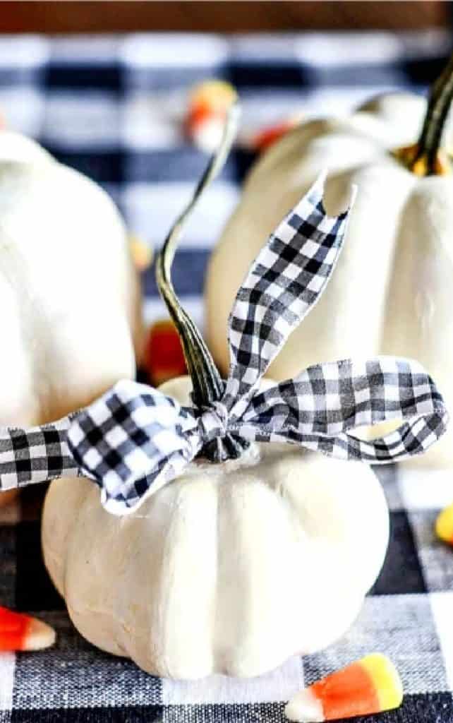 White faux pumpkin tied with a black and white gingham ribbon bow on a buffalo check cloth with candy corn, a simple idea for decorating with faux pumpkins.