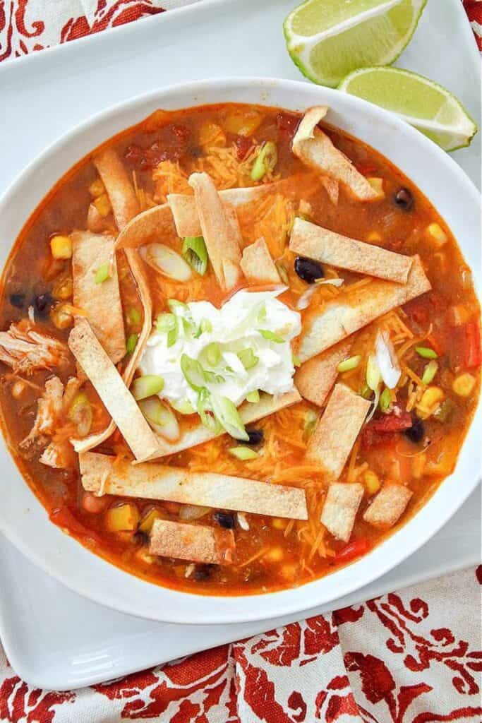 Chicken taco soup in a white bowl topped with tortilla strips, sour cream, shredded cheese, and green onions, with lime wedges on the side.