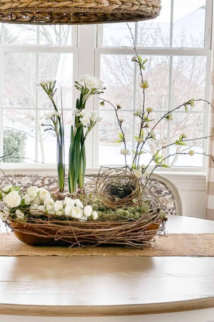 Early spring dough bowl arrangement with blooming branches, paperwhite bulbs, ranunculus, moss, grapevine, and a nest on a dining table.