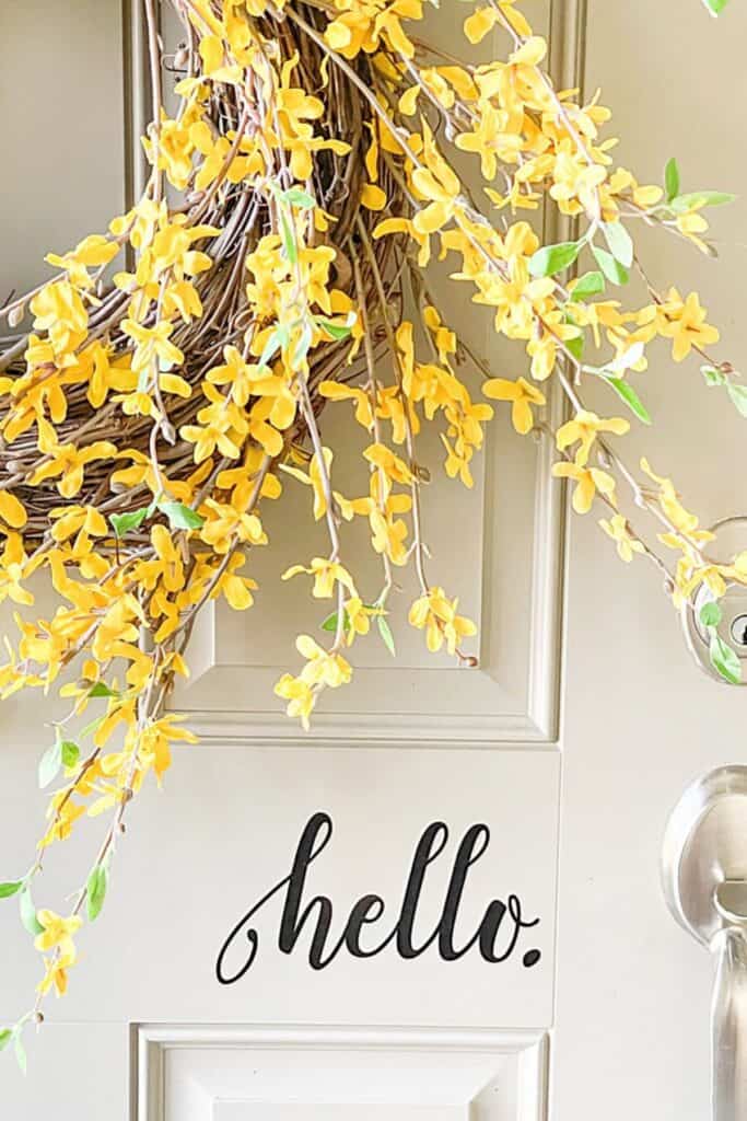 Yellow forsythia wreath with trailing branches hanging on a light colored front door with a hello sign.