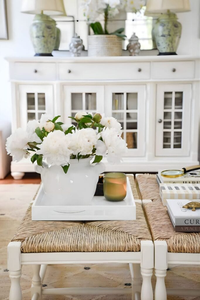 Layering decor on a coffee table with a white tray, peonies, candle, and books in a living room