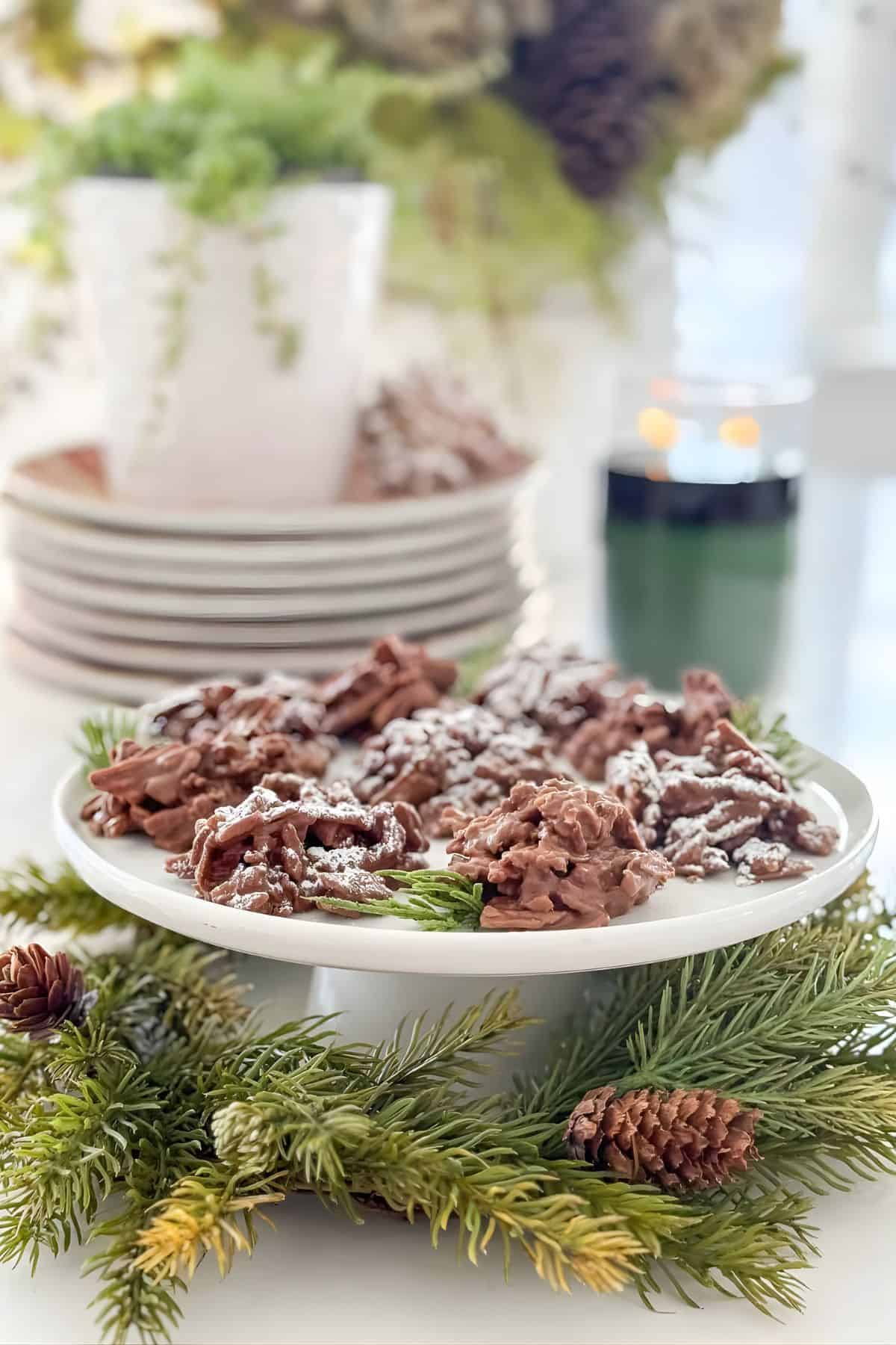 No bake haystack cookies dusted with powdered sugar on a white cake stand with Christmas greens