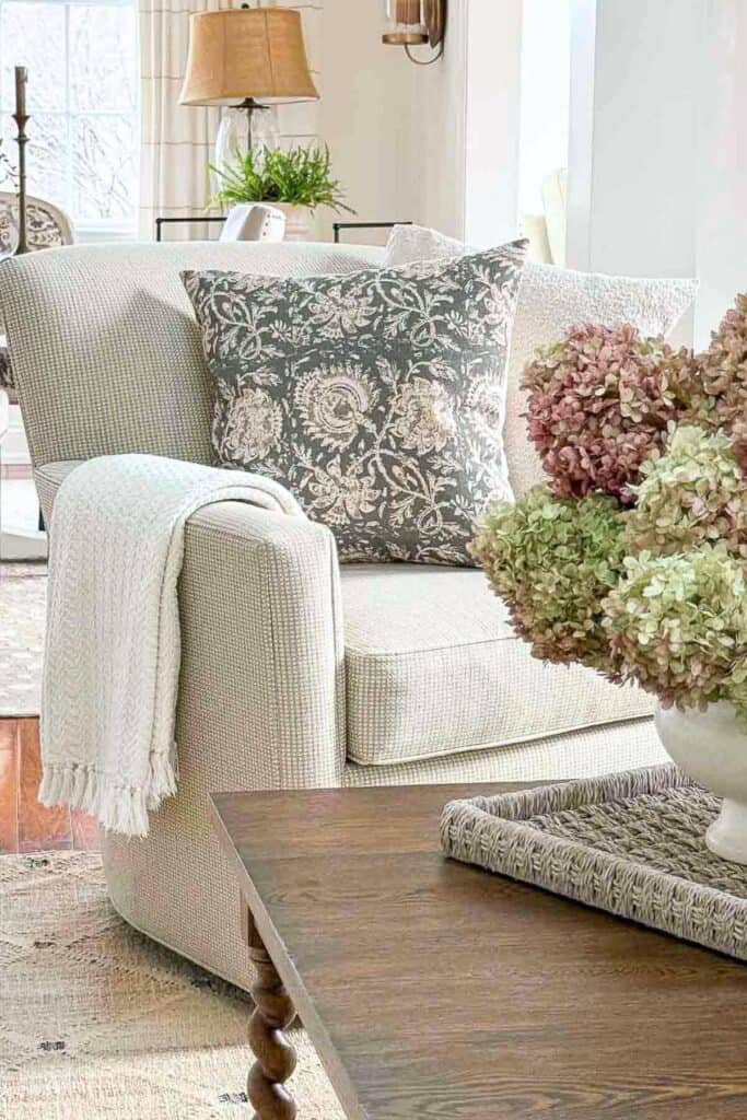 Upholstered living room chair styled with a patterned pillow, a light throw draped over the arm, and a coffee table with a woven tray and flowers nearby