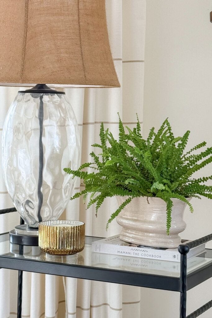Side table styled with a glass table lamp, a potted fern set on a book, and a small gold candle beside it