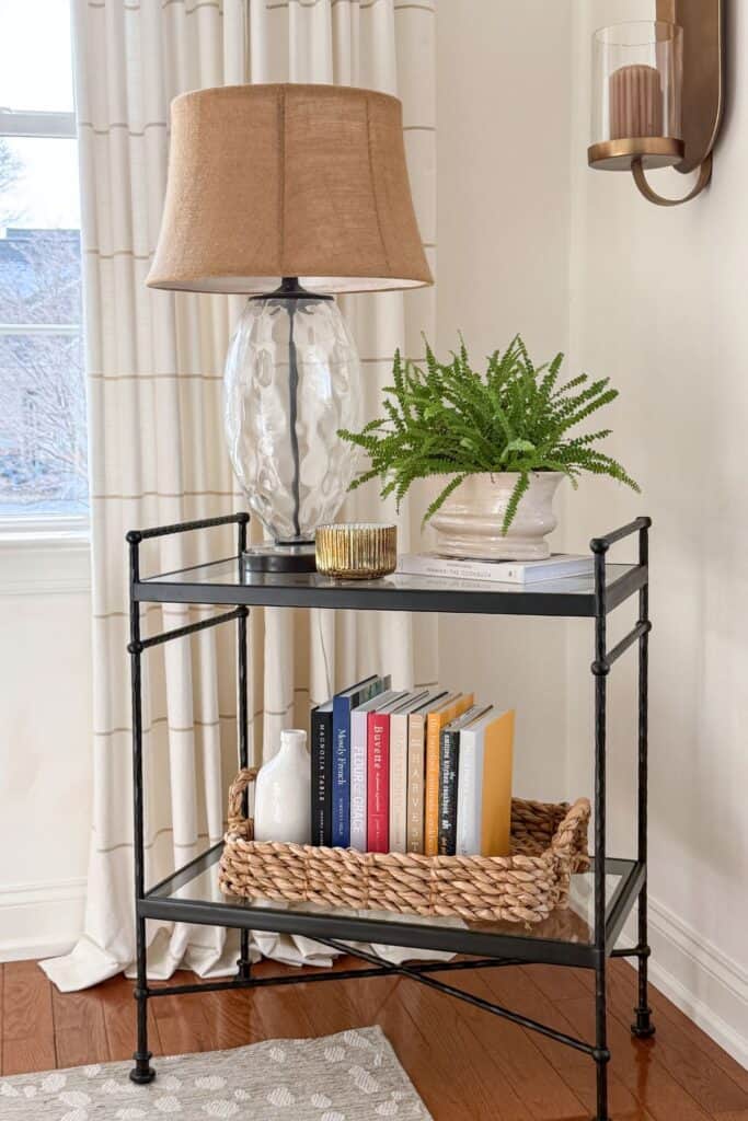 Side table styled with a glass table lamp, a potted fern set on a book, and a small gold candle beside it