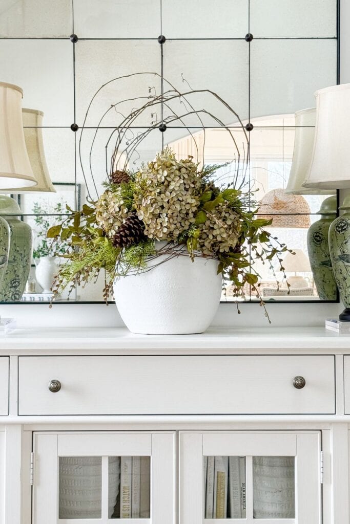 White buffet styled with a large urn filled with greenery and branches, flanked by matching table lamps and a paneled mirror above