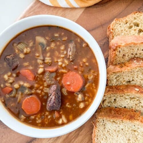 Bowl of homemade beef barley soup with carrots and tender beef served with sliced bread on a wooden board.
