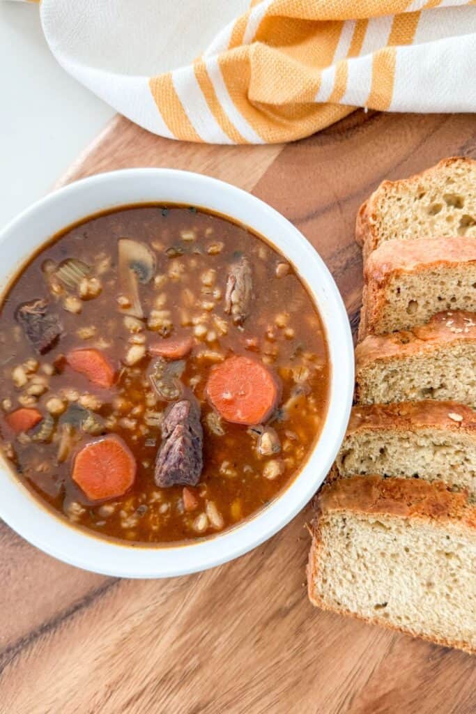 Bowl of homemade beef barley soup with carrots and tender beef served with sliced bread on a wooden board.