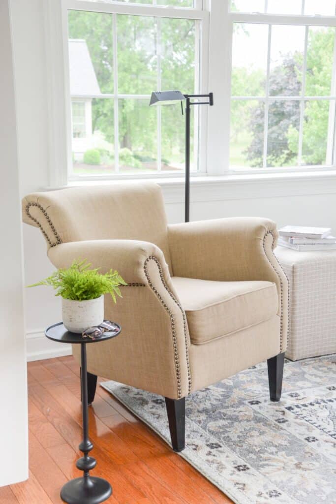 Neutral upholstered armchair with nailhead trim, small round side table, and floor reading lamp placed near large windows in a bright living room.