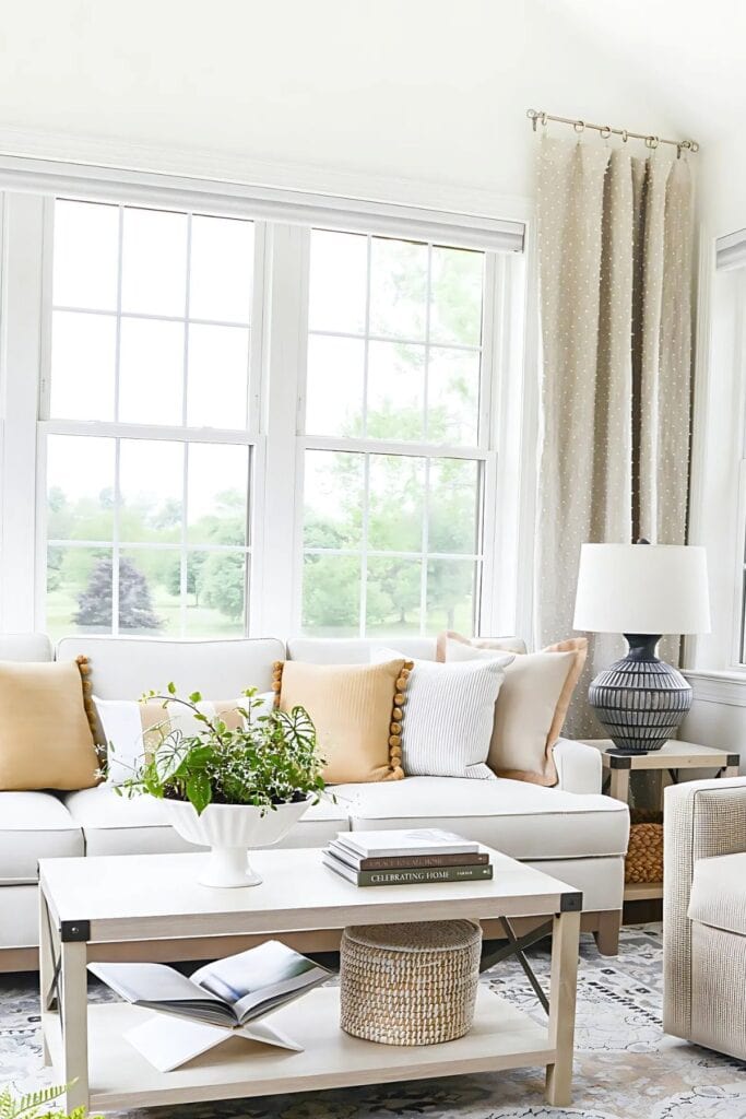 Neutral living room with white sofa, layered pillows, and coffee table styled with books and greenery in a bright sitting area.