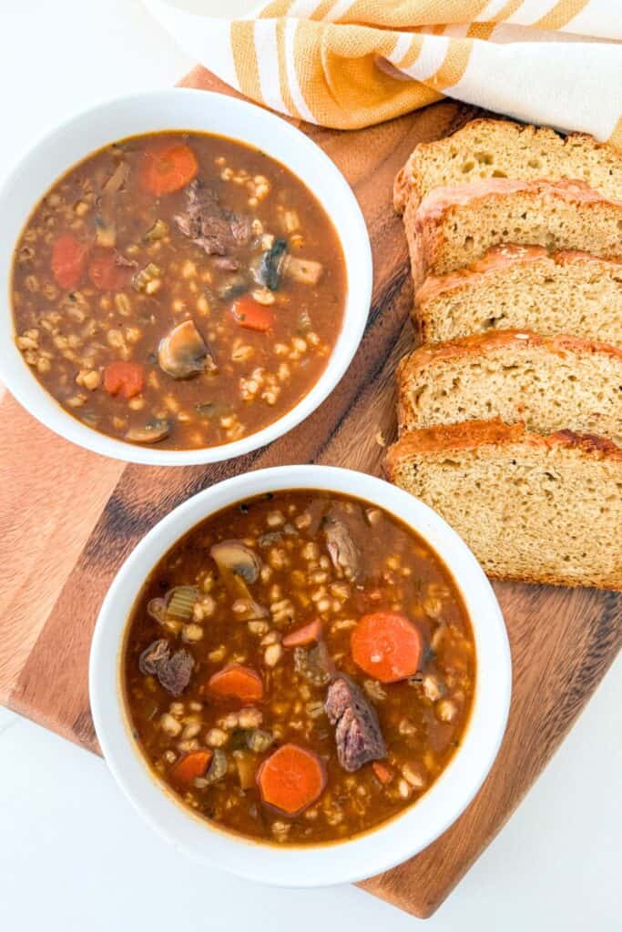 Two bowls of homemade beef barley soup with carrots, pearl barley, and tender beef served with sliced bread on a wooden board.