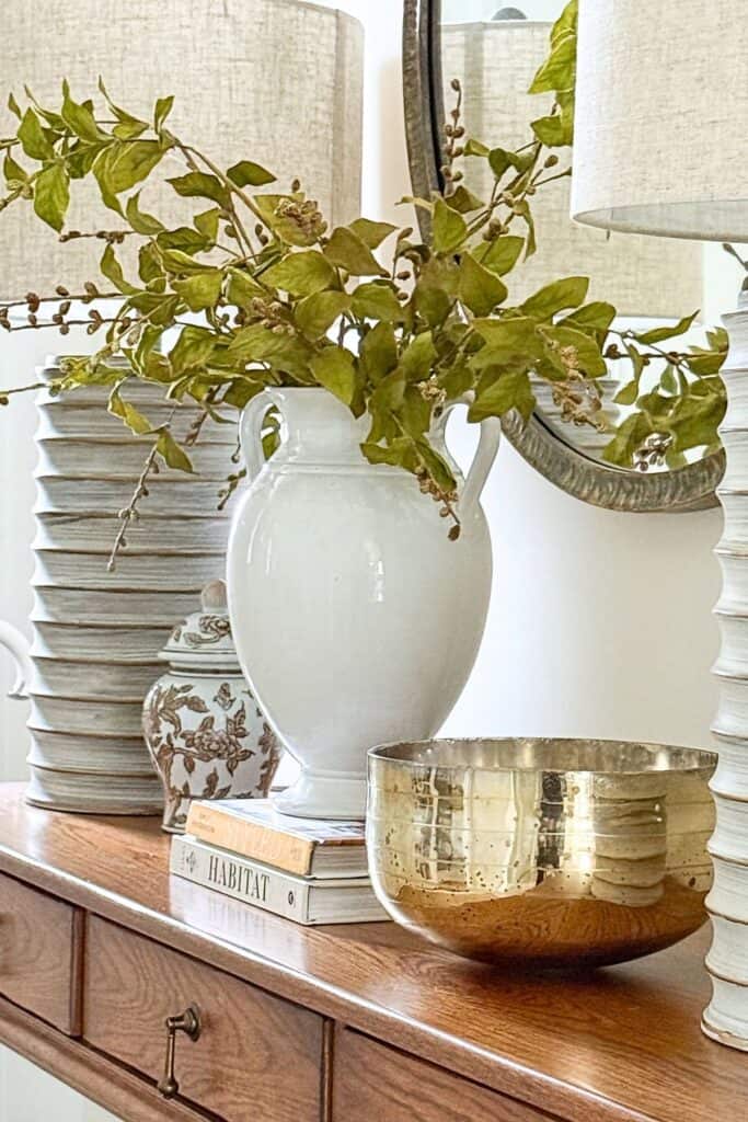 Collected console table decor with layered lamps, white vase with greenery, brass bowl, and stacked books styled in a balanced, clutter free way.