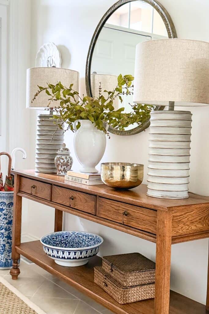 wood console table styled with white lamps, greenery in vase, round mirror, gold bowl, and layered decor for a collected look without clutter