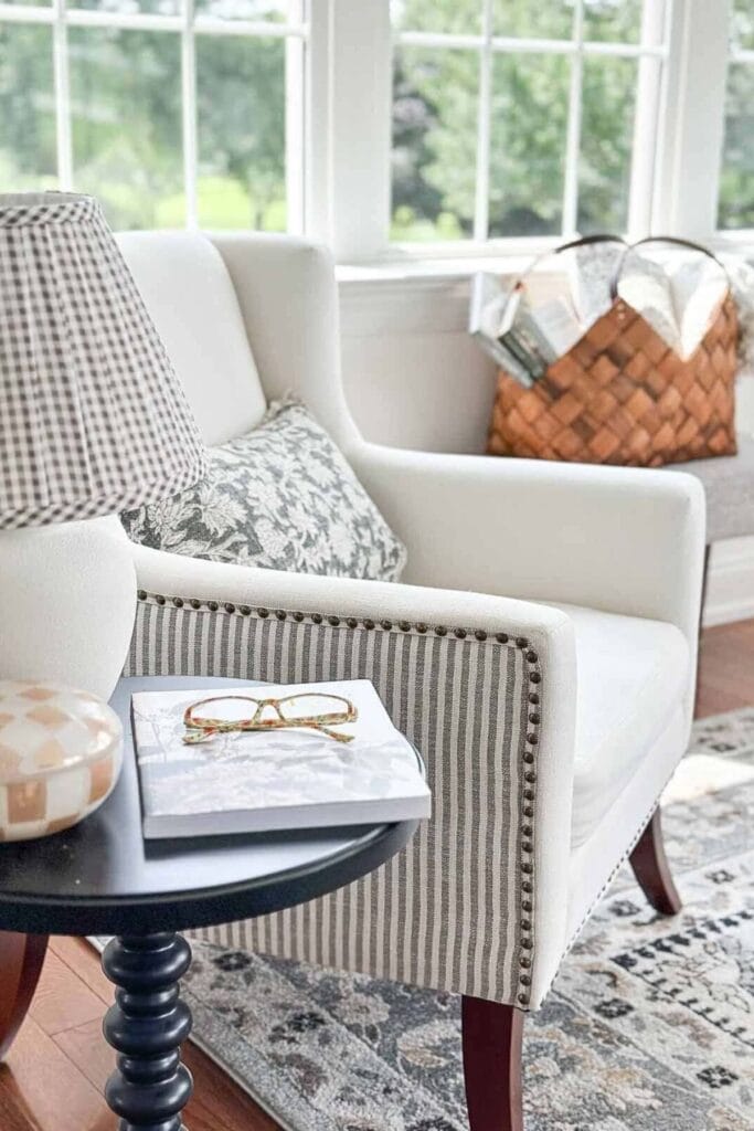 Neutral armchair with striped trim and patterned pillows, styled side table with book and glasses, and woven basket in a bright sitting area.