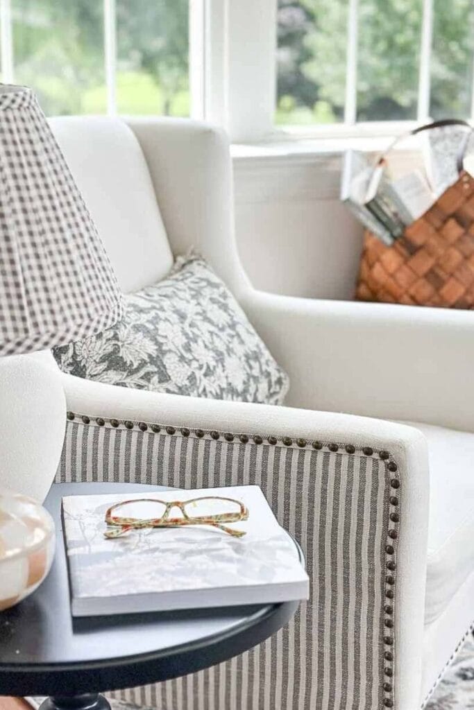 Neutral striped armchair with nailhead trim, patterned pillows, and a side table styled with a book and reading glasses in a light filled sitting area.